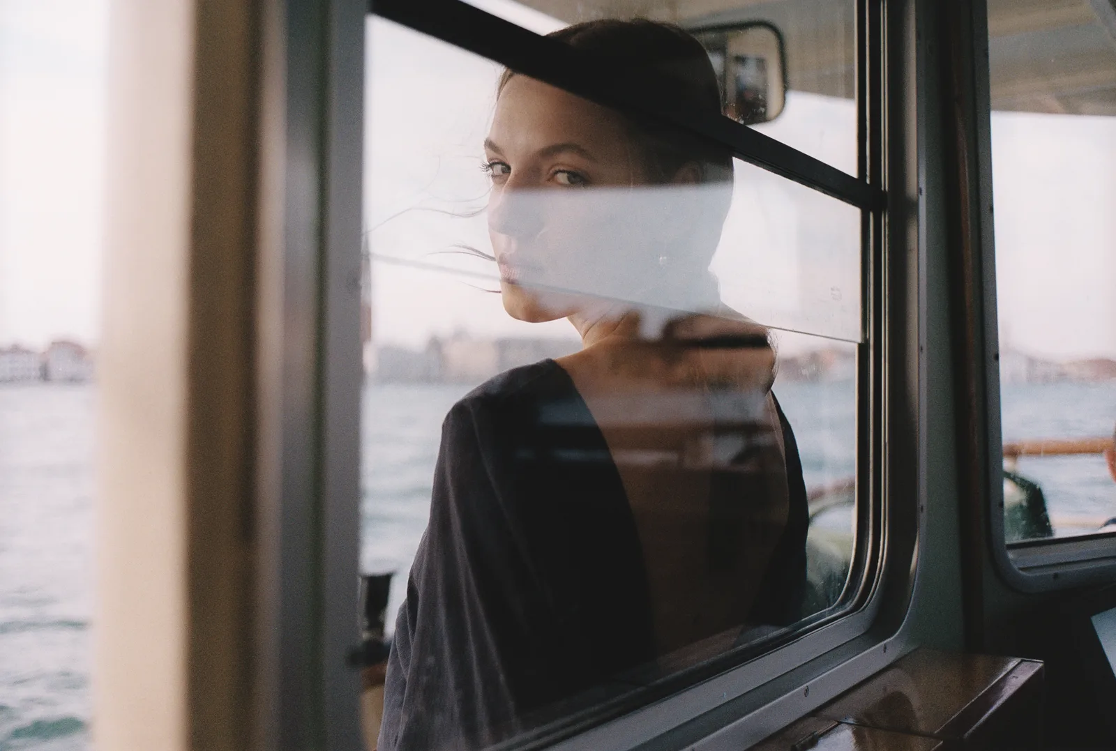 A woman on a vaporetto looking back through the window, the Venice lagoon and the far shore beyond her, reflections lying across the glass — a colour negative developed by Trichroma Lab.