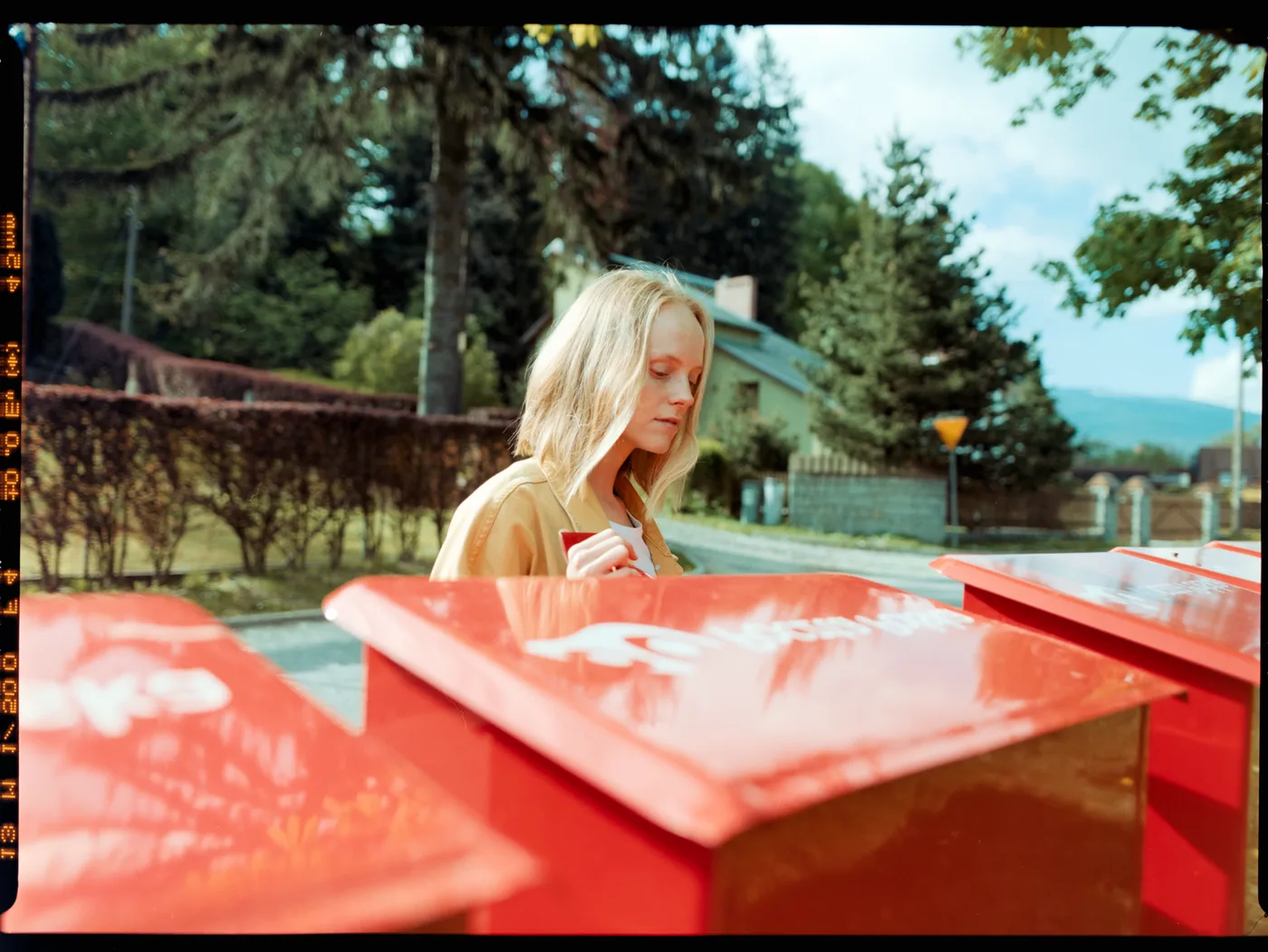A blonde girl in a mustard jacket behind three red boxes, trees and a house behind her, on Portra 160, converted by Negative Lab Pro from a white-light raw.