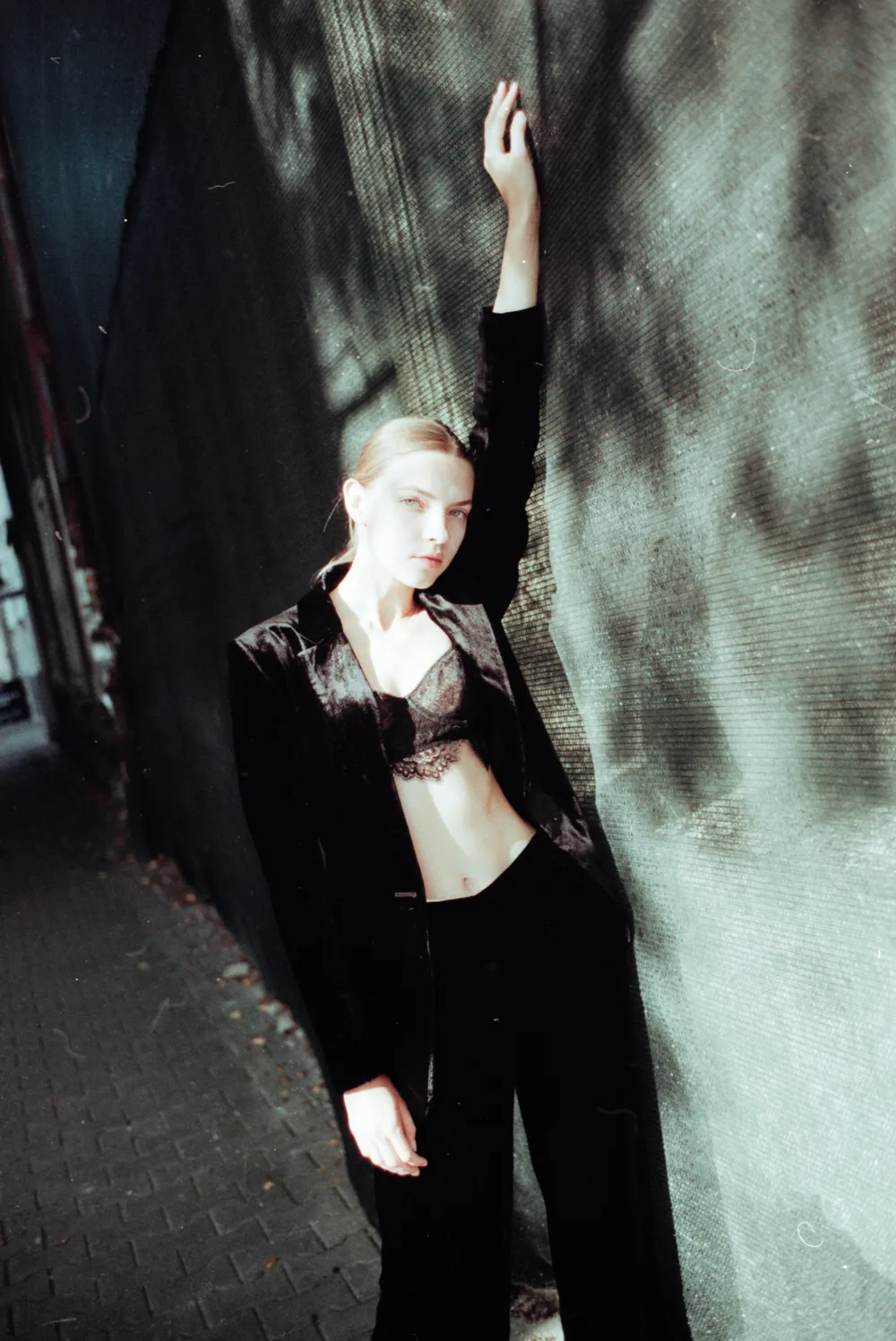 A woman in a black velvet suit leaning on a wall in dappled shade, one arm raised, on Gold 200, an Epson V700 scan converted by Negative Lab Pro.