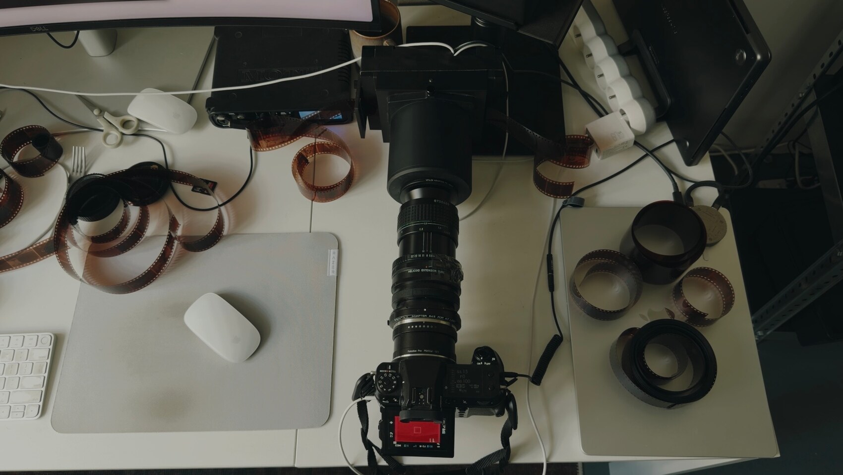 The copy stand from above: the camera and its long lens pointing down at the film holder, with a strip of negatives running through the gate and more film on the desk around it.