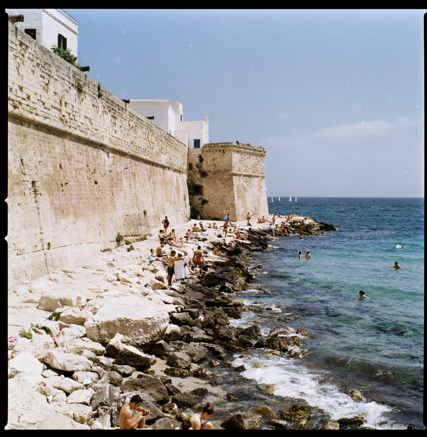 A tall stone sea wall above a rocky shore, bathers on the rocks and in clear turquoise water, white houses on top.