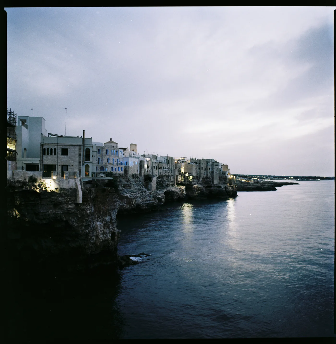 A town of white houses on a cliff above the sea at dusk, the first lights reflected in calm water.