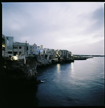 A town of white houses on a cliff above the sea at dusk, the first lights reflected in calm water.