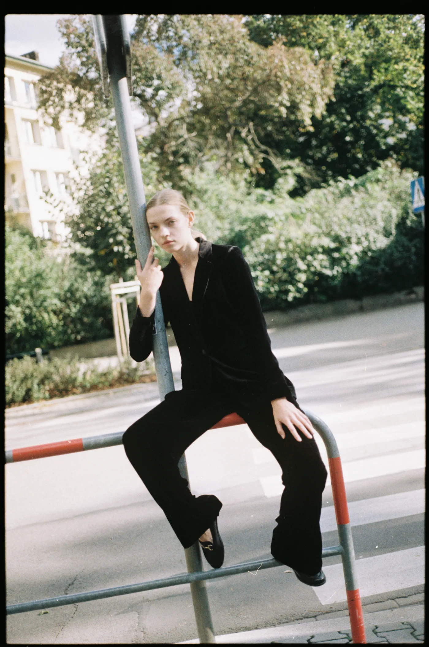 A young woman in a black velvet suit sitting on a red-and-white street railing beside a pole.