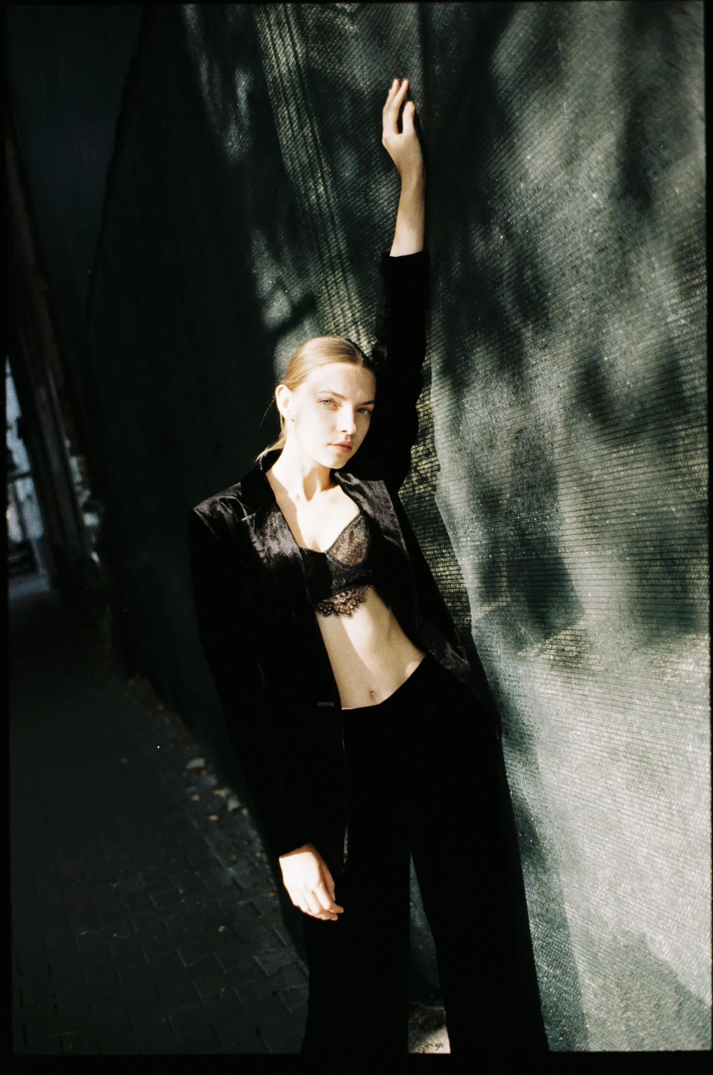 A young woman in a black velvet suit leaning against a shadowed wall, one arm raised.