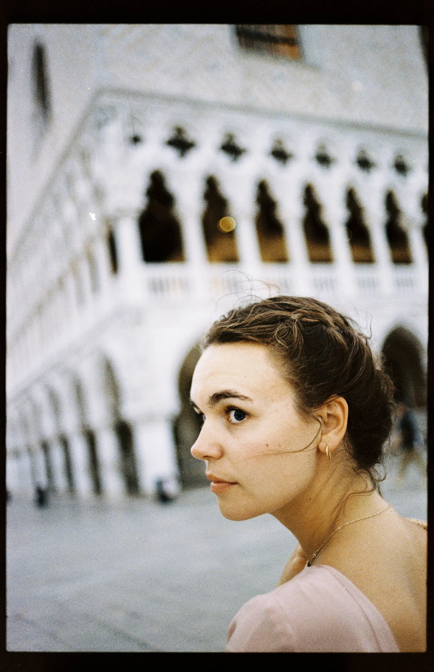 A young woman with her hair pinned up looks over her shoulder, a white Gothic arcade blurred behind her.