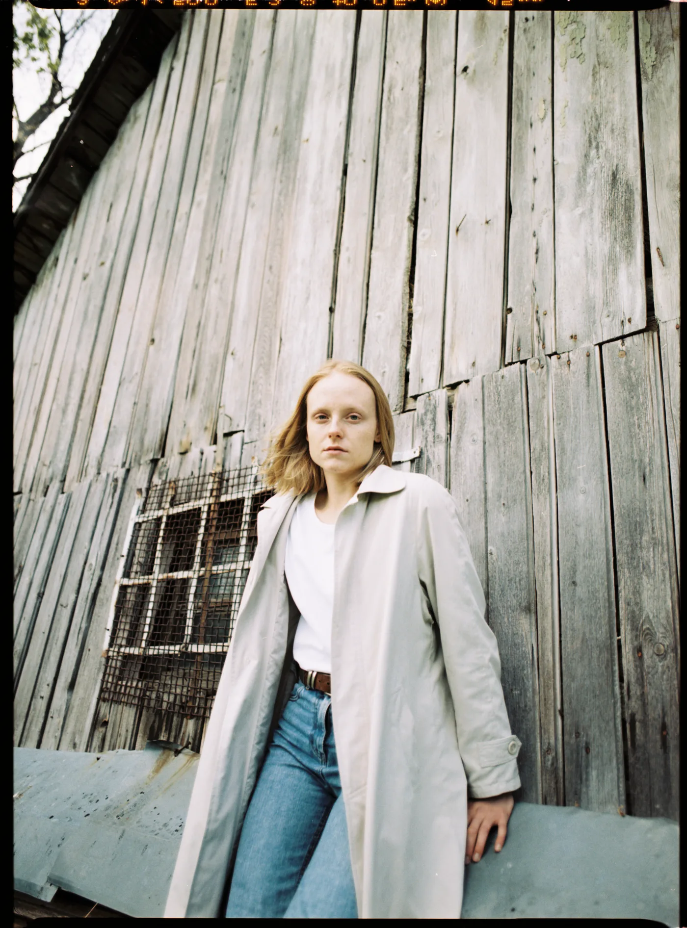 A woman in a pale trench coat leaning against the grey boards of an old wooden barn.