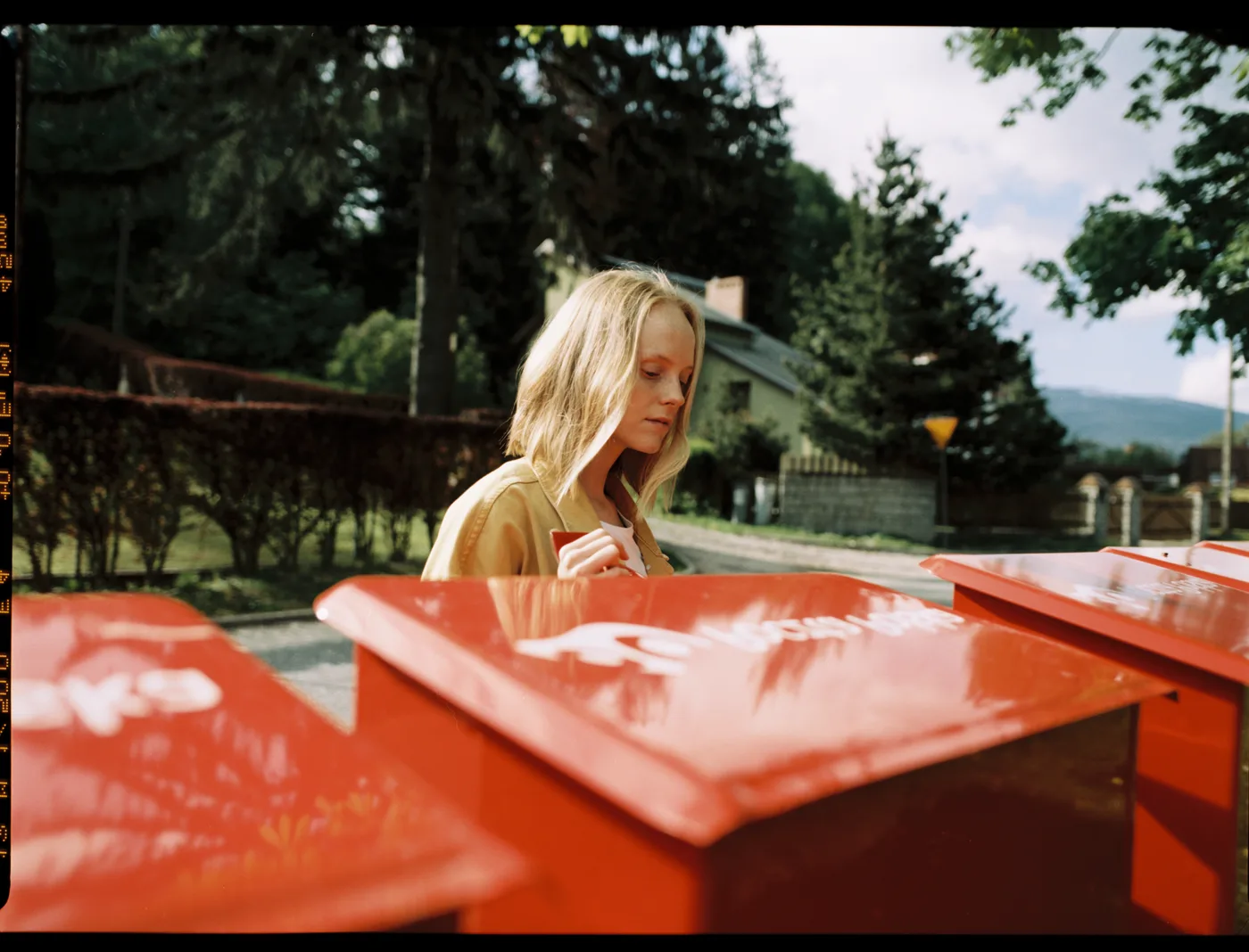 A blonde woman in a mustard jacket behind a row of red post boxes, pines and a house behind her.