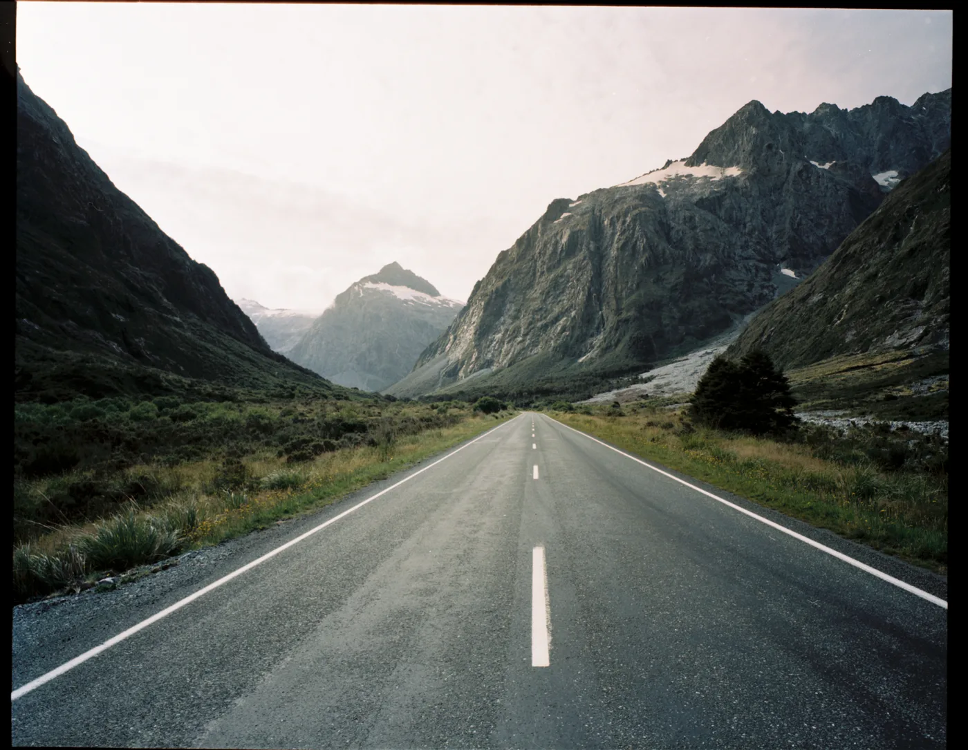 An empty road running straight into a valley between steep mountains with patches of snow.