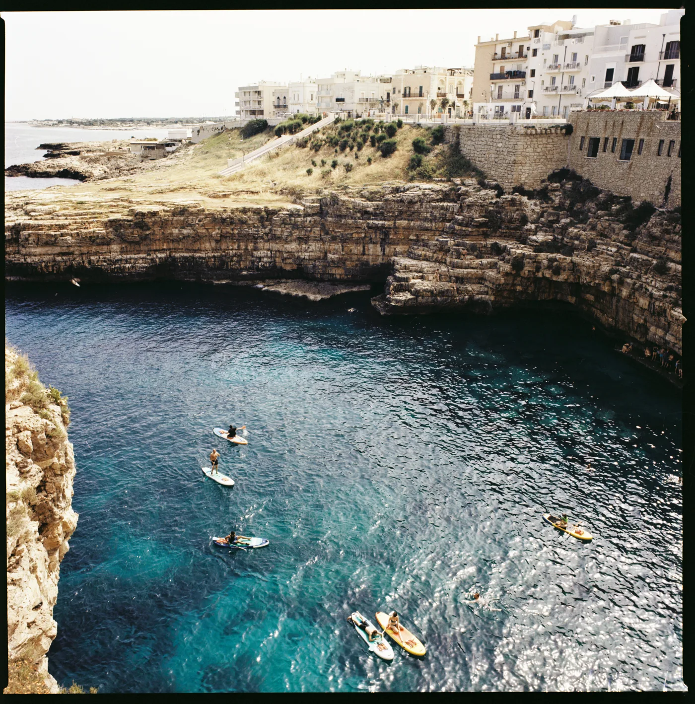 Paddleboarders on deep blue water in a cove ringed by cliffs and white houses.