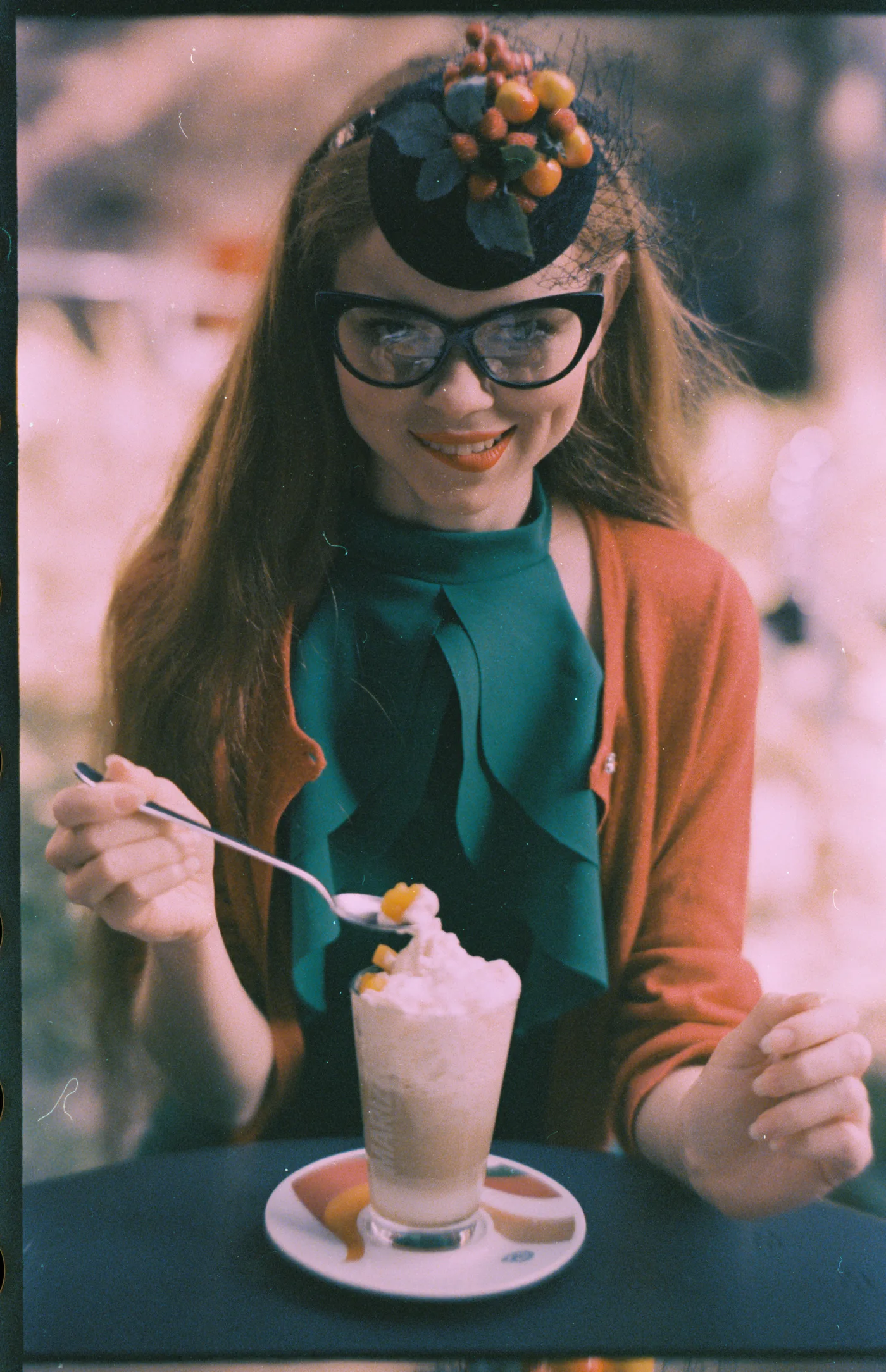A smiling woman in cat-eye glasses and a hat trimmed with berries, eating whipped cream from a tall glass.
