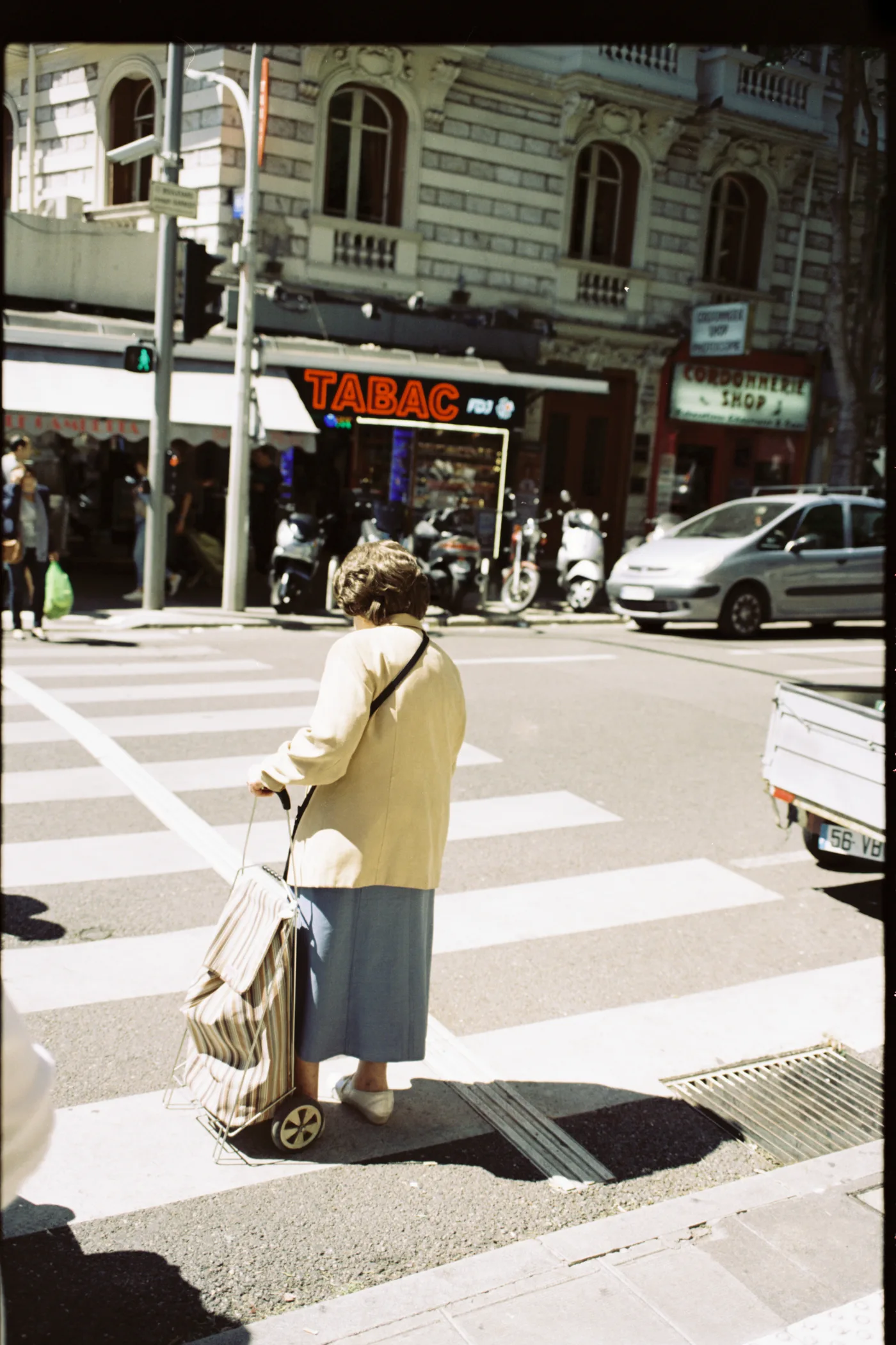 An elderly woman with a shopping trolley waiting at a pedestrian crossing, a tabac behind her.